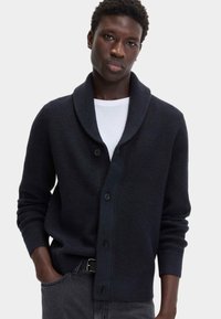 Marineblauwe gebreide cardigan met een sjaalkraag, knopen aan de voorkant en geribbelde manchetten. Gecombineerd met een wit T-shirt en donkere jeans.