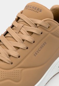 Bruna sneakers med texturerad gummiyta, beige snören och perforerade detaljer. Har "SKECHERS" branding och en vit sula.