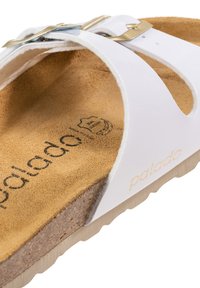 Sandal blanc avec une semelle intérieure texturée marron, dotée d'une boucle dorée et d'un détail de logo. Le matériau semble lisse et contemporain dans son design.