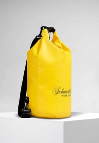 Sac étanche jaune avec une fermeture enroulable, sangle réglable noire et logo imprimé en noir. Texture lisse, forme cylindrique.