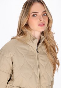 Beige gesteppte Jacke mit steigendem Kragen, mit Reißverschluss und strukturiertem, welligem Muster auf dem Stoff.