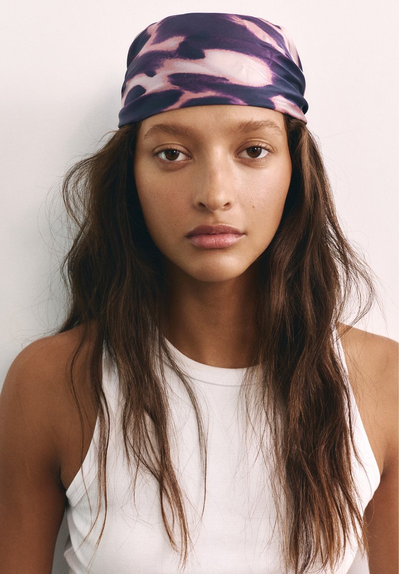 Jeune femme aux longs cheveux bruns portant un foulard violet à motifs et un haut blanc sans manches, regardant vers l’avant avec une expression neutre.