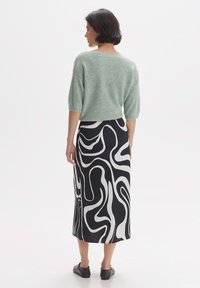 Lichtgroene katoenen trui met korte mouwen, gecombineerd met een zwarte midi rok met een wit abstract golvenpatroon en zwarte platte schoenen.