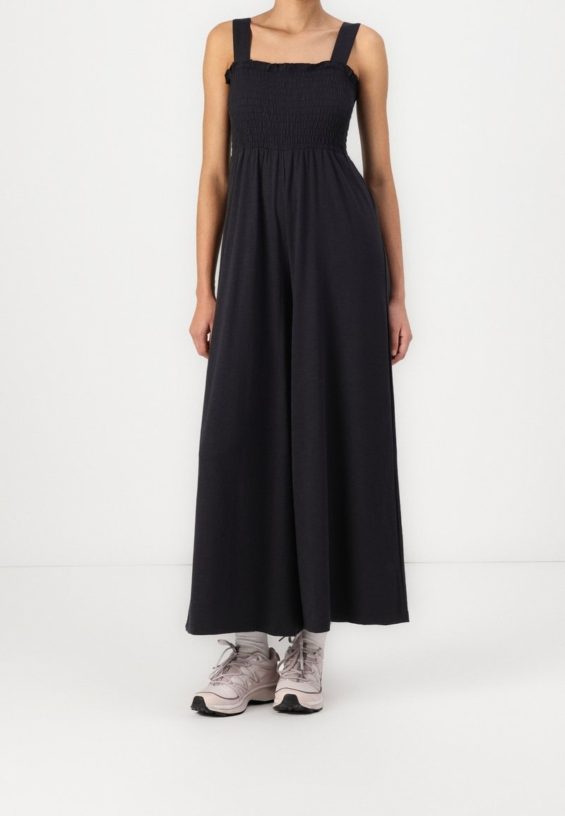 Femme portant une combinaison noire sans manches à jambes larges avec un corsage smocké et des baskets roses avec des chaussettes blanches sur fond blanc.