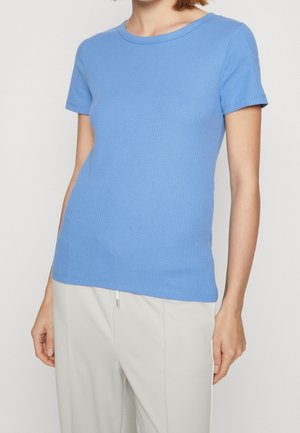 Kobieta ubrana w jasnoniebieski, prążkowany, krótki sweter z krótkim rękawem, w połączeniu z jasnobeżowymi spodniami, pokazana od szyi do bioder na jednolitym tle.