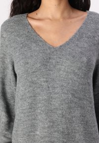 Pull en V gris fabriqué dans un tissu doux et texturé. Dispose d'une coupe décontractée et de manches longues, avec des coutures visibles le long de l'encolure.