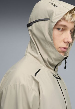 Beige leichte Jacke mit Kapuze, ausgestattet mit einem schwarzen Reißverschluss, verstellbarem Kordelzug und Logoakzenten. Glatte Textur mit minimalen Designdetails.