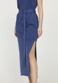 Femme portant une jupe midi bleue ceinturée avec une fente latérale haute, montrant sa jambe nue et du vernis à ongles rouge sur sa main.