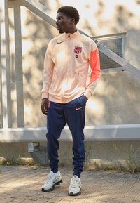 Nike track jacket in pale peach with pink accents and logo, paired with navy sweatpants and white athletic shoes. Textured fabric throughout.