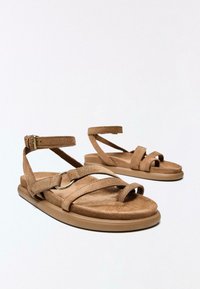 Sandales plates en daim marron avec brides à la cheville, boucles pour les orteils et détail d’anneau métallique, présentées sur un fond blanc uni.