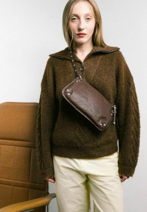 Maglione a trecce marrone con colletto zip abbinato a pantaloni color crema. Una borsa a tracolla in pelle marrone scuro presenta dettagli in metallo.