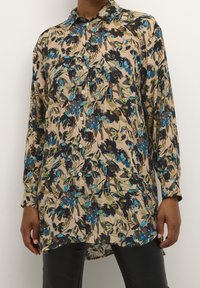 Chemise à boutons à imprimé floral beige avec des accents bleus, verts et noirs. Manches longues, col, et détail de l'ourlet arrière plus long.