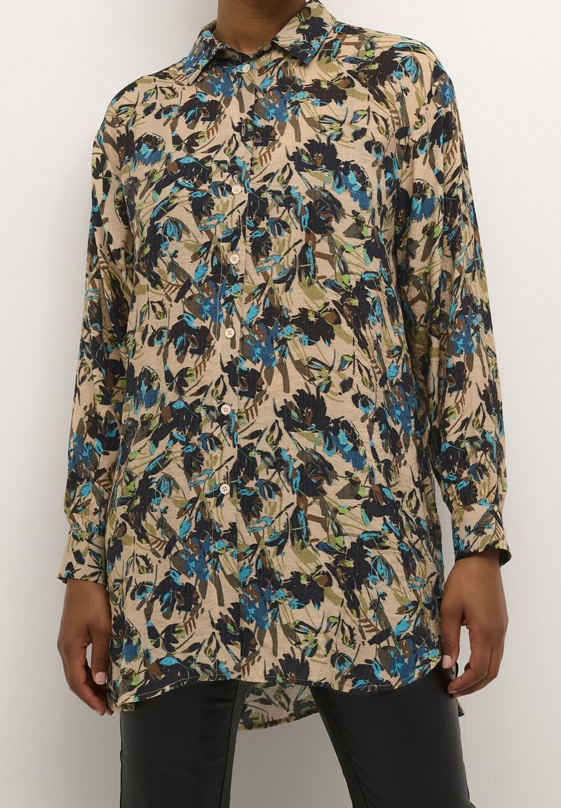 Chemise à boutons à imprimé floral beige avec des accents bleus, verts et noirs. Manches longues, col, et détail de l'ourlet arrière plus long.