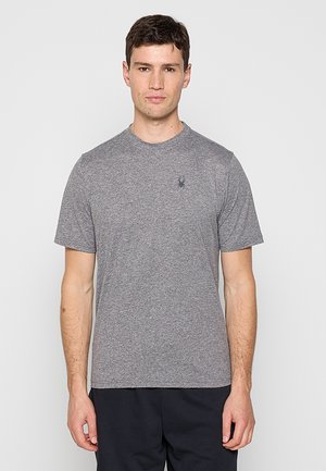 Mężczyzna o krótkich ciemnych włosach, ubrany w gładką szarą koszulkę z małym czarnym logo pająka na lewej piersi, stoi na tle jednolitego jasnego tła.