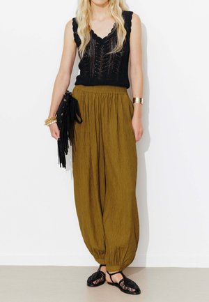Femme blonde portant un haut en maille noir sans manches, pantalon ample vert olive, ballerines noires tissées, tenant une pochette noire à franges avec des bracelets aux poignets.