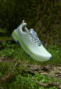 Ανοιχτό πράσινο και μπλε παπούτσι για τρέξιμο σε μονοπάτι που αιωρείται πάνω από το δάπεδο ενός δάσους με ορατό γρασίδι, κλαδιά και χώμα από κάτω.
