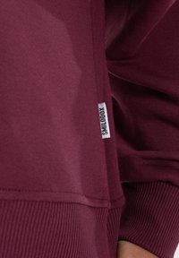 Sweatshirt bordeaux avec des poignets et un ourlet côtelés, doté d'une petite étiquette blanche portant "SMILLODOX" sur le côté. Tissu doux et texturé.