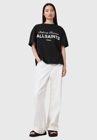 T-shirt oversize nera con testo bianco "Infinite Summer ALLSAINTS LONDON." Abbinata a pantaloni bianchi a gamba larga e sandali neri.