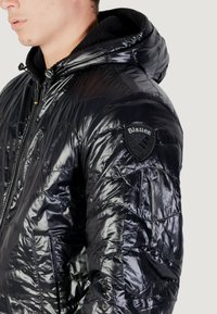 Giacca piumino nera con un finish lucido e trapuntato. Presenta un cappuccio, chiusura con zip e una toppa con "Blauer" sulla spalla sinistra.