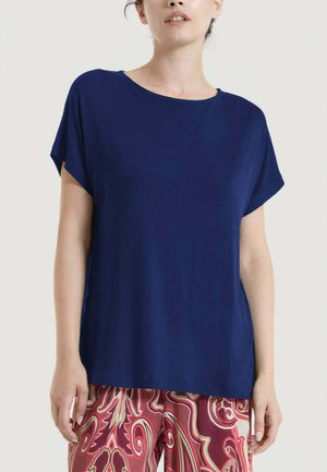 Donna che indossa una maglietta blu navy a maniche corte dal taglio ampio e pantaloni larghi a fantasia rossa, rosa e beige, in piedi contro uno sfondo uniforme.