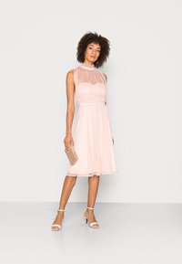 Vestido em rosa claro, sem mangas, com um decote alto transparente e uma cintura definida, saia fluida, combinado com sapatos de salto brancos com tiras e uma clutch dourada.