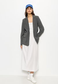 Grauer Blazer, weißes Maxikleid, weiße Sneaker und blaue Mütze. Der Blazer hat ein maßgeschneidertes Design mit einer einzelnen Tasche.