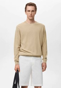 Beige long-sleeve sweater with a textured finish, paired with white shorts, and held with a black bag. Simple design, no visible patterns.