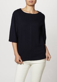 Maglione blu navy a maglia cable con scollatura arrotondata e maniche a tre quarti, realizzato in tessuto testurizzato. Indossato sopra pantaloni chiari.