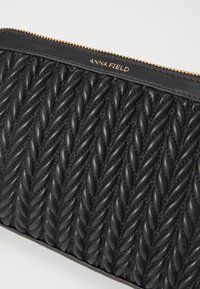 Clutch de cuero negro con una textura similar a cuerda. Presenta accesorios en dorado y un cierre superior. Nombre de la marca "ANNA FIELD" grabado en la parte frontal.