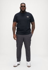 Tricou negru cu mâneci scurte și un mic logo pe față, asortat cu pantaloni cargo gri închis și adidași albi cu dungi negre.