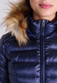 Doudoune marine avec fermeture éclair et bordure de capuche en fausse fourrure. Le tissu est lisse et brillant, avec des motifs matelassés horizontaux.