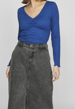 Femme portant un haut bleu à manches longues et col en V, ainsi qu'une jupe en jean gris foncé taille haute avec une fente à l'avant.