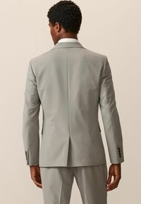Traje gris claro con una sola abertura, corte entallado y puños abotonados. Textura de tela suave y diseño minimalista.