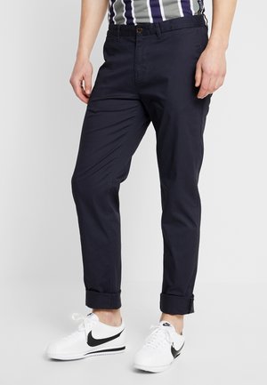 Mężczyzna noszący ciemnogranatowe podwinięte spodnie chino połączone z białymi butami Nike z czarnym logo Swoosh na jasnym jednolitym tle.