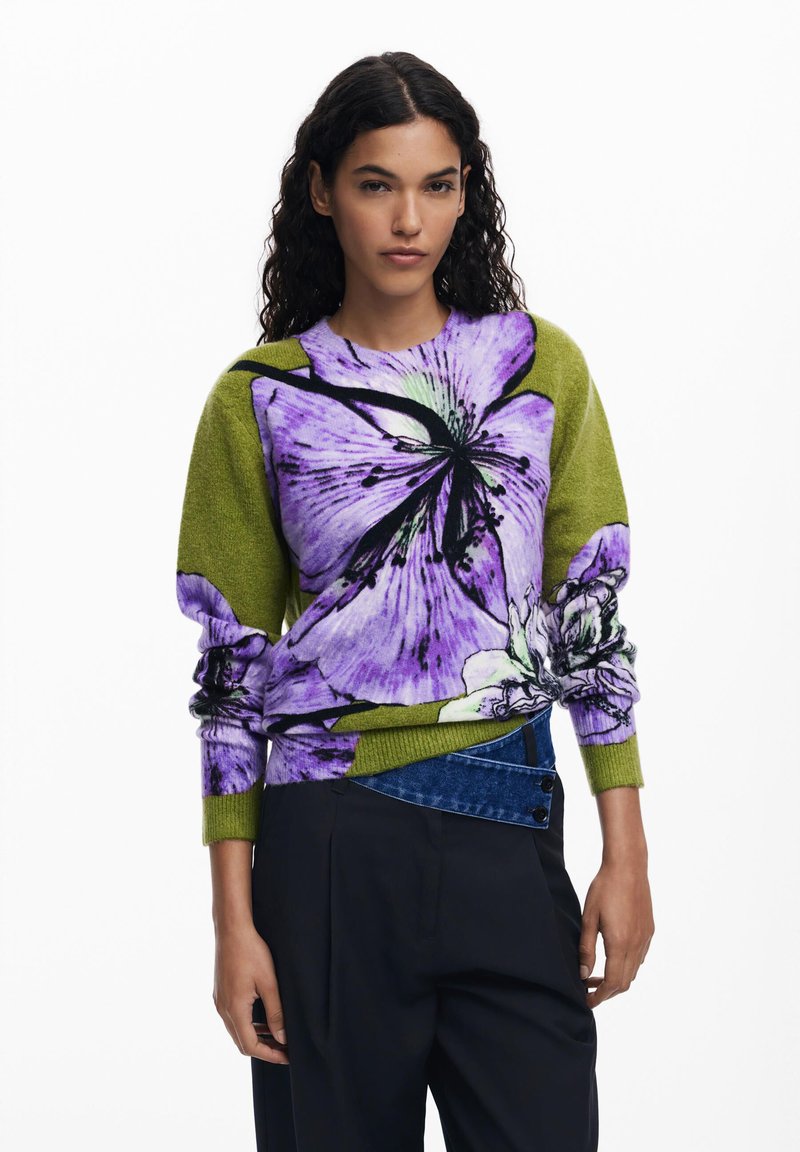 Knit sweater featuring a vibrant purple floral design on a green background, with ribbed cuffs and a relaxed fit. Paired with dark trousers.