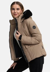Beige pufferjack met een zwart bont-achtige capuchon, ritssluiting aan de voorkant en een logo patch. Heeft zijzakken en geribbelde boorden voor extra warmte.