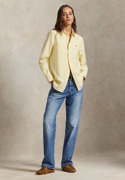 Frau steht mit Händen in den Taschen, trägt ein blassgelbes Hemd mit Knöpfen, blaue Jeans, schwarzen Gürtel und braune Mokassins.