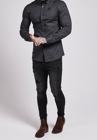 Camisa negra de botones hecha de una tela con un ligero brillo, combinada con jeans negros desgastados y botas de ante marrón hasta el tobillo.