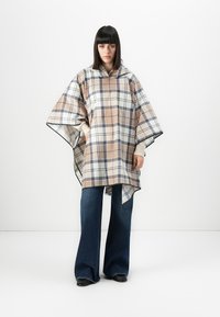 Woman with straight dark hair wearing beige and white plaid poncho with hood over cream sweater and wide-leg dark blue jeans against white background.