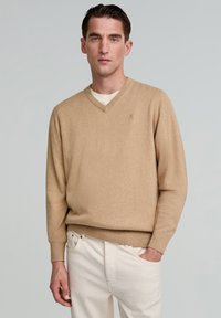 Beżowy sweter w serek wykonany z miękkiego materiału dzianinowego, z długim rękawem i ściągaczami, noszony na jasnej koszulce, w połączeniu z jasnymi spodniami.