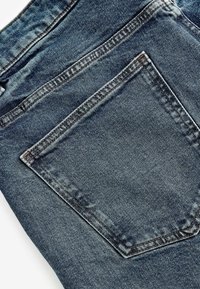 Material denim de culoare bleu închis, evidențiind un buzunar din spate cu cusături vizibile. Detalii subtile de uzură și textură sunt evidente pe întreaga suprafață.