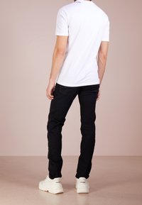 T-shirt bianca a maniche corte con dettagli neri, abbinata a jeans neri slim-fit e sneakers bianche chunky. Vista posteriore su uno sfondo neutro.