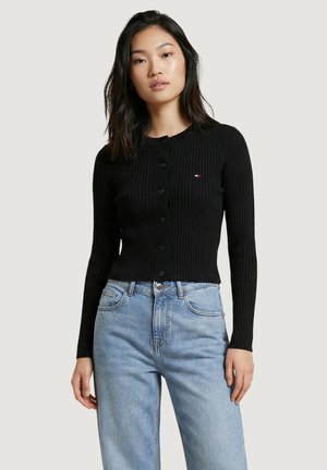 Giovane donna con capelli neri di lunghezza media, che indossa un maglione nero a costine con bottoni e jeans a vita alta di colore azzurro chiaro.