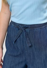 Top albastru cu dungi, asortat cu pantaloni de denim închis, cu talie ajustabilă cu șnur și țesătură texturată. Șnurul este legat într-o fundă.
