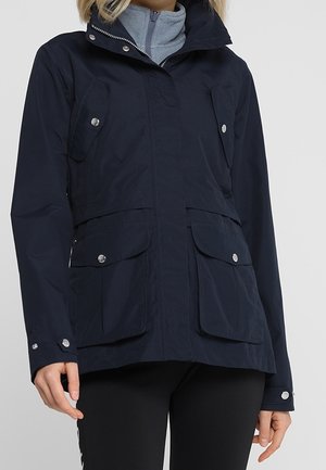 Femme portant une veste bleu marine avec fermeture éclair à l’avant et quatre poches à rabat avec boutons-pression argentés, associée à un pantalon noir.