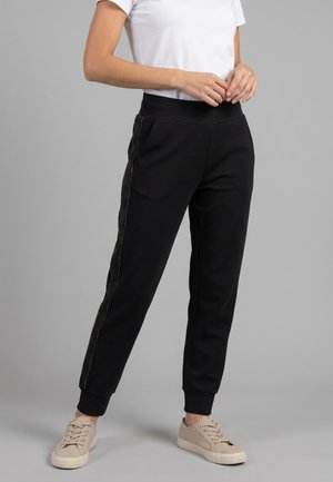 Personne portant un pantalon de jogging noir avec un détail de bande latérale, un t-shirt blanc et des baskets beiges, debout sur un fond gris.