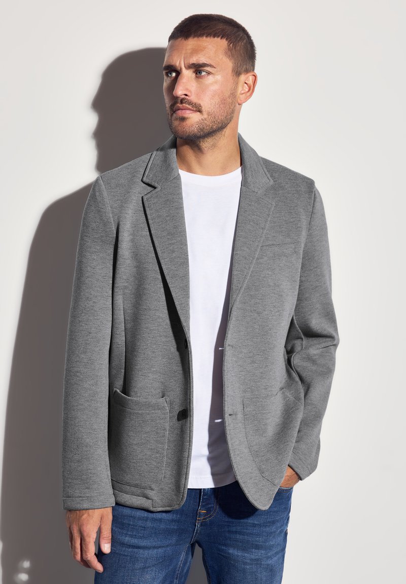 Harmaa bleiseri, joka on valmistettu pehmeästä kankaasta ja jossa on lovettu kaulus, kaksi etutaskua ja rento malli, käytetty valkoisen T-paidan ja sinisten farkkujen päällä.