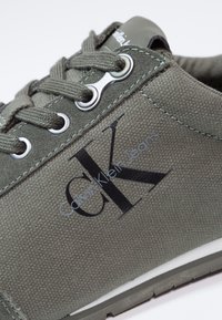 Gröna tygskor med grå snörning, ögla och en svart logo "CK" med texten "Calvin Klein Jeans". Gummisula med vita accenter.