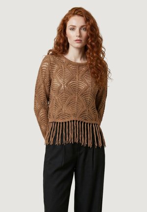 Donna con lunghi capelli rossi ricci, indossa un maglione marrone lavorato a uncinetto con frange e pantaloni neri larghi, in piedi davanti a uno sfondo semplice.