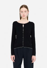 Mujer llevando un cárdigan negro con ribetes plateados y botones decorativos, combinado con pantalones negros y pendientes largos plateados, de pie contra un fondo blanco.